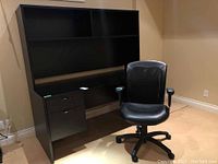 Full frontal view of black desk with shelving and drawers alongside adjustable black office chair with visible armrest tear.