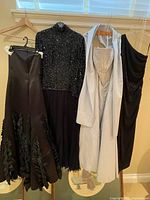 Photo of four evening gowns hanging on hangers side by side on a rack beneath a window.