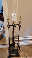 Set of four fireplace tools with black shafts and brass handles hanging from black metal stand with brass finials and trim, shown against cream wall and hardwood floor.