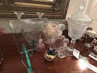 Photo showing a collection of glass decorative items including footed lidded dish, pedestal bowl, glass basket, various glass decanters, and a green-tinted glass display stand with etched glass plates.
