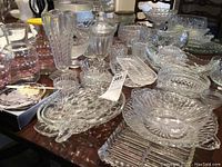 Full view of assorted clear glass serving pieces on a table including bowls, footed bowls, relish dishes, juice jugs, and covered jar.