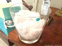Clear glass ice bucket filled with tissue paper. Round handle on side. Tiffany & Co. sticker visible.