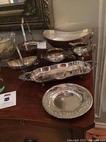 Photo showing multiple silver plated items including tray, cream pitcher, sugar bowl, decorative dishes on a wooden table.