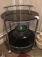 Full view of glass-topped round table with chrome frame and curved rail on back, with an art glass bowl on lower shelf.