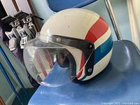 Vintage red, white, and blue open-face helmet with clear visor placed on a chair, skis and poles in the background.