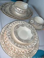 Display of various plates, bowls, cups, and serving pieces showing gold floral pattern on cream base