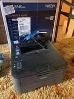Photo showing Brother HL L2340DW printer with power cord in front of original box on carpeted floor.