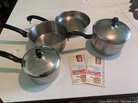 Five Farberware stainless steel pots with lids and black handles, plus one unmarked pan without lid.