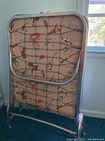 Metal folding cot frame standing upright showing underside with bungee-style cord support and western themed mattress fabric.