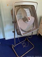 Photo showing the full fan including the metal frame, adjustable stand, and three large metal blades behind a protective metal grille, with the Fasco logo in center