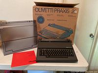 Olivetti Praxis 20 typewriter placed in front of its original cardboard box with the plastic cover and instruction manual visible.