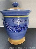 Blue ceramic tea jar with floral vine design and 'Tea of Courage' text, showing lid with yellow knob.