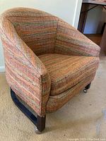 Front side view showing barrel-shaped chair with upholstery in multi-colored orange, green, and brown fabric with wooden base and casters.