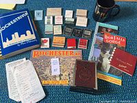 Photo showing Rochester NY memorabilia including books, assorted matchbooks, maps, a brown mug, and various paper items.