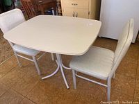 Photo of small rectangular kitchen table with cream formica top and 2 padded chairs with worn white vinyl upholstery and pastel print