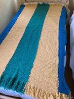Full view of the vintage handmade afghan blanket spread out on a bed, showing the five stripes in blue, yellow, and teal with fringe on both ends.
