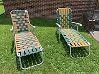 Two vintage lounge lawn chairs with green and gold webbing on aluminum frames placed on grass in front of brick wall