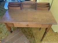 Full view of solid wood writing desk showing top surface with two raised organizer compartments and single drawer, standing on turned spindle legs.