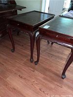 Two antique Victorian wooden tea tables with rectangular tops and cabriole legs shown on a wooden floor. Visible slide-out shelves and small drawer with brass knob.
