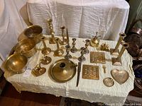 Full shot of brass decor items spread out on white cloth, showing pots, bowls, candle sticks, trays, small bell, and decorative brass pieces.