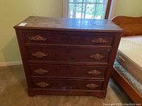 Front view of wooden chest with four drawers and decorative carved handles. Surface shows wear and scratches.