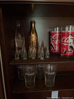 Shelf displaying various Coca-Cola glasses and miniature bottles, including two tall plastic cups with vintage red labels.