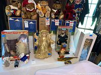 Four dolls display on table, including porcelain angel doll in gold, three boxed porcelain dolls, three small stuffed bears, and two vintage toys in front of blue boxes of teddy bears in background.