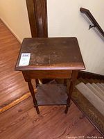 View of the wooden end table from an angle showing top surface, legs, and lower shelf near stairway.