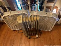 Two white Lesko box fans placed upright with visible control dials and an oil-filled radiator heater between them on a wooden floor.