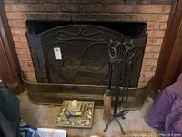 Black metal fireplace screen with ornate scrollwork, brass mesh fireplace screen, fireplace tool stand with broom, poker, and shovel, small plug-in fountain with rocks and shells on hearth.