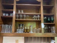 Assorted vintage glassware, including stemmed glasses, short tumblers, amber-colored glasses, decorative wine bottles, and candleholders arranged on wooden shelves.
