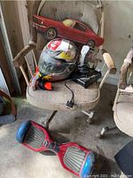 Red hoverboard on floor in front of chair. Motorcycle helmet with flames and Nintendo Zapper gun on chair seat. Joystick next to helmet.