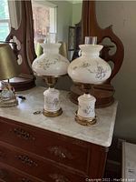 Pair of two early 1900s white ceramic lamps with purple and yellow floral hand-painted designs, brass fittings, and glass chimneys, displayed on a wooden dresser with mirror.