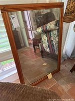 Front angled view of large rectangular mirror with wooden frame, reflecting a window and furniture.