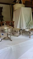 Three silver-tone chafing dishes and food holders displayed on a white cloth-covered table showing the set as a whole.