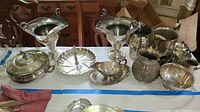 Photo showing a large assortment of silver plate serving pieces including large handled covered dishes, pitchers, bowls, and trays on a white tablecloth.