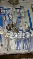 Multiple assorted silver plated forks, spoons, and serving utensils, some in plastic bags with labels.