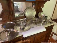 Full display of various sterling silver dishware pieces on wooden sideboard showing multiple trays, cups, and serving bowls with visible tarnish, needing cleaning.