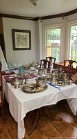 Full view of a large wooden table with a white tablecloth and silverware on top in a dining room with wood flooring.