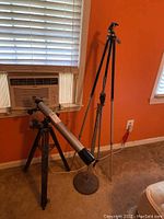 Bushnell telescope mounted on a black tripod next to a silver mic stand in a room with orange walls and carpeted floor.