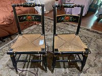 Pair of vintage black wooden chairs with floral hand painted detailing on the back and woven rush seats, showing overall wear including seat wear and surface scratches.