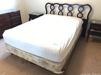 Full view of bed with white mattress, floral box spring, intricate wood headboard and metal frame on casters in bedroom.