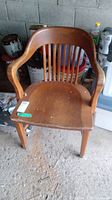 Front view of vintage oak chair showing curved backrest with vertical slats and armrests. Chair seat shows surface wear.