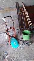 Photo showing the Whirl-on spreader, blue lawn dolly, and wooden garden hand tools against a brick wall.