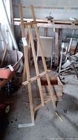 Wooden easel shown standing upright with adjustable shelf visible, inside a workshop space.