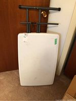 Folding utility table folded stored against a door on carpeted floor, showing black metal legs and white table top.