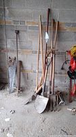 Nine assorted garden tools including shovels, pitchforks, hedge shears, sprinkler, saw, and auger leaning against wall