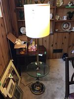 Metal floor lamp with off-white fabric shade, clear round glass table, metal base and pole, photographed in a wood-paneled basement room near a desk and other lamps.