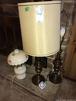 Three vintage table lamps: two tall metal base lamps (one with beige fabric shade, one without) and one smaller white ceramic, glass, metal hurricane lamp plus a lamp timer on floor in basement near fireplace