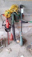 Black and Decker leaf blower, hedge trimmer, grass trimmer alongside a yellow extension cord against a brick wall.